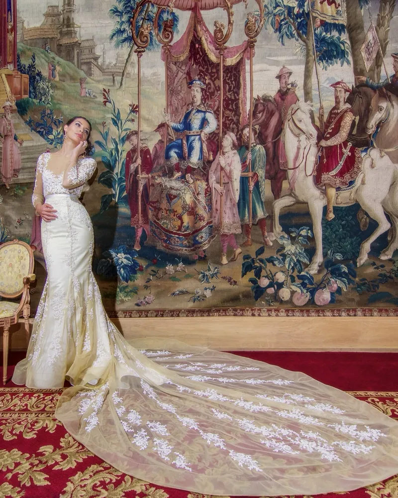 Euterpe. A bride in a lace couture gown with a sweeping train, in front of a tapestry.